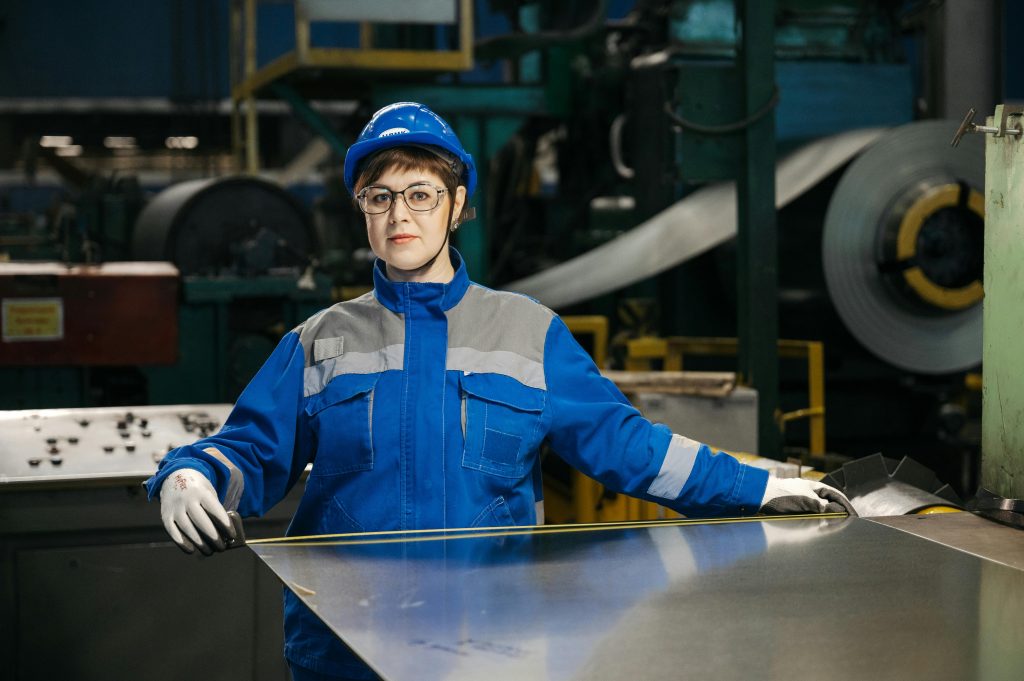 Female factory worker in protective gear handling a metal sheet in an industrial setting. Safety and industry focus.